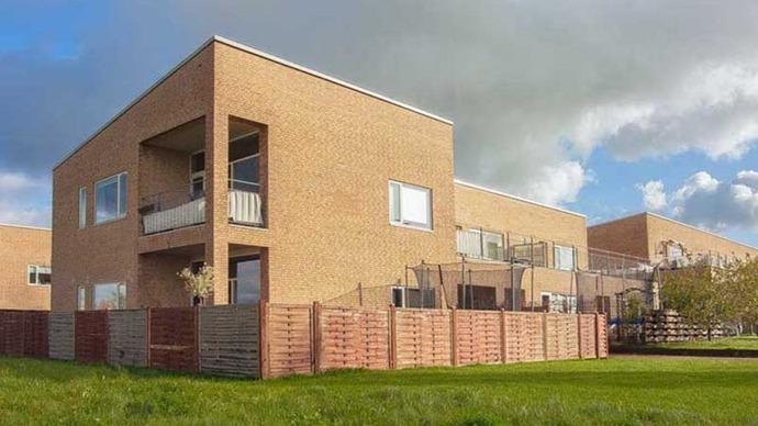 Moderne murstenshus med balkon og græsplæne, omgivet af et hegn under en skyet himmel.
