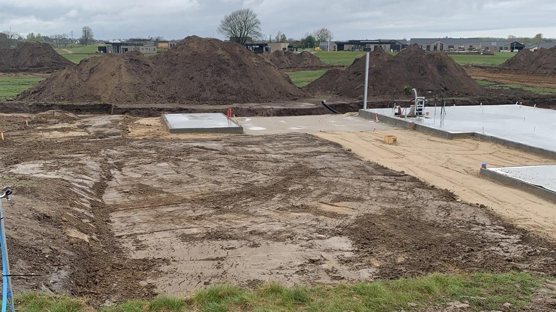 Byggeplads med udgravet grund, sand og betonplader, omgivet af jordhøje og græsarealer.