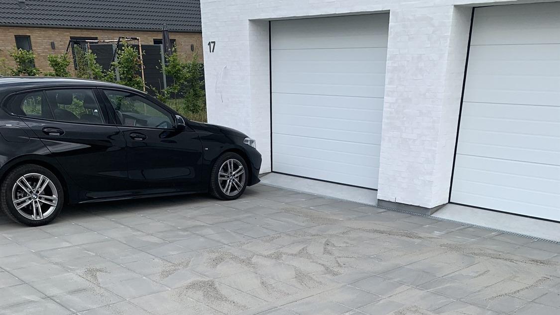 En sort bil parkeret ved siden af to garagedøre på en flisebelagt indkørsel.