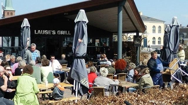 En travl restaurant med mange gæster, der sidder ved borde udenfor, under parasoller på en solrig dag.