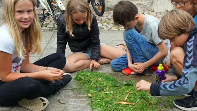 Børn sidder på jorden og laver kunst med græs og blomster, mens de koncentrerer sig om deres kreative projekt.