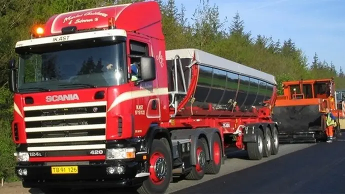 En rød Scania lastbil med en stor trailer parkeret ved vejen, omgivet af grønne træer.