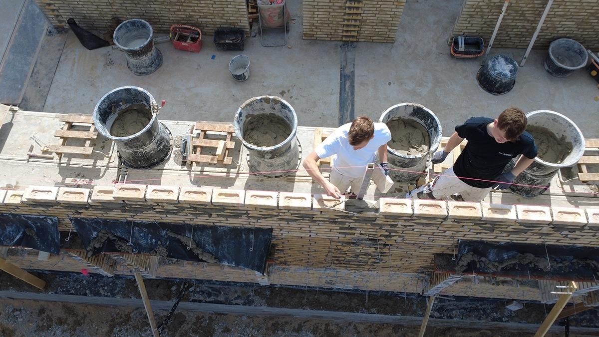 To personer, der arbejder på en murbygning, med cementspande og mursten i baggrunden.