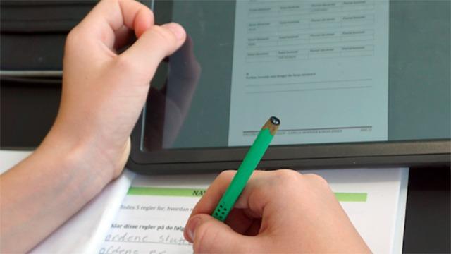En hånd holder en grøn blyant, mens den anden hånd bruger en tablet til at skrive på et papir med opgaver.