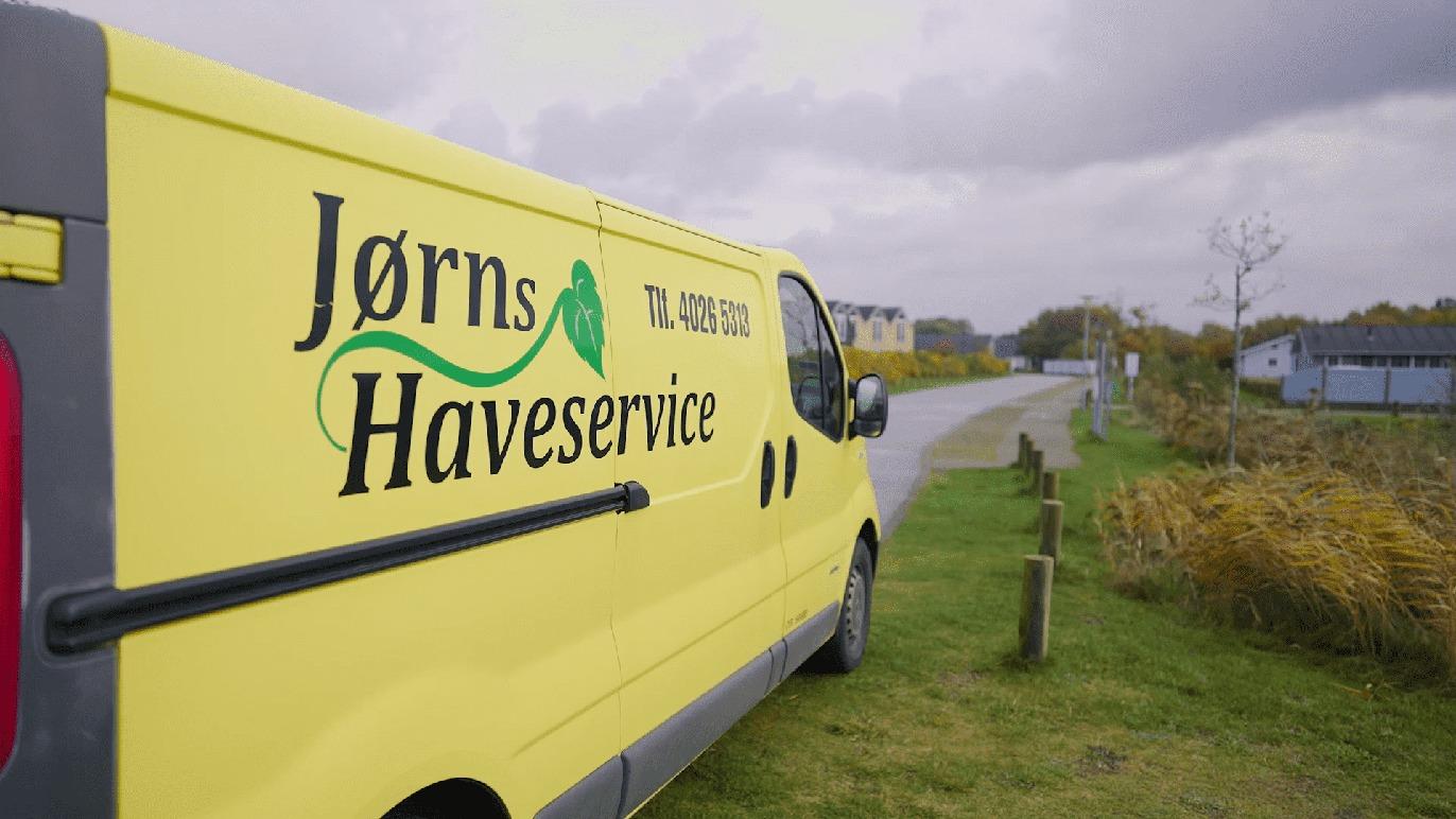 Gul varevogn fra Jørns Haveservice parkeret ved en vej i et landskab med græs og træer.