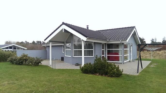 Et gråt hus med skråtag og store vinduer, omgivet af grøn græsplæne og buske.