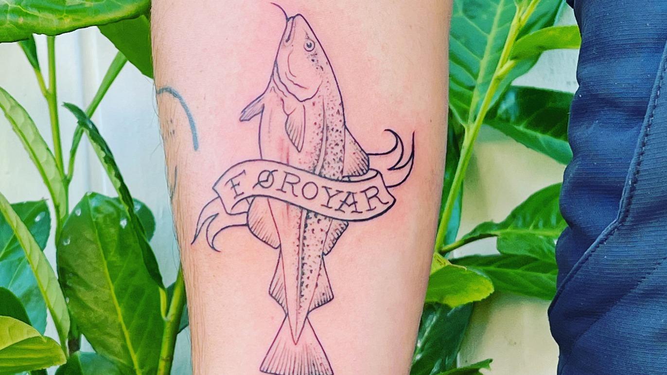Tatovering af en fisk med teksten "F. Øroyar" på en arm, omgivet af grønne planter.