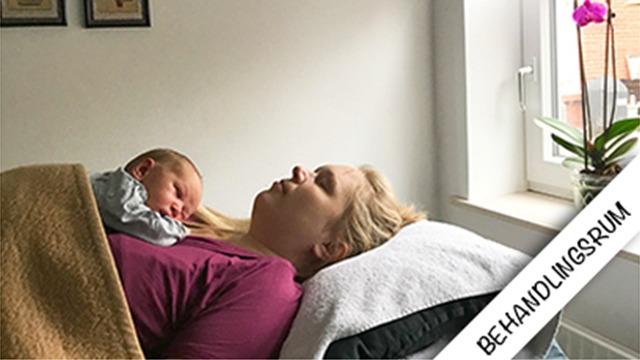 Mor ligger på en behandlingsbriks med en sovende baby på sit bryst, omgivet af et lyst rum med vindue og plante.