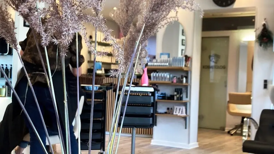 En frisørsalon med pampasgræs i forgrunden og en frisør, der arbejder i baggrunden.