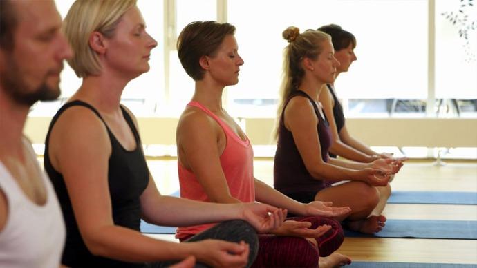 Gruppe af mennesker, der mediterer i en yogaklasse, siddende på måtter med lukkede øjne og afslappede ansigtsudtryk.