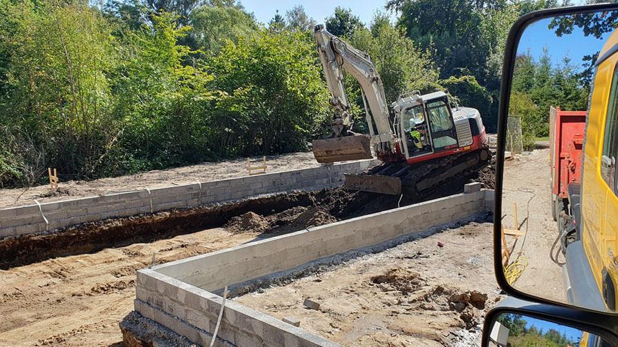 Gravemaskine arbejder på en byggeplads med betonfundamenter og grønne træer i baggrunden.