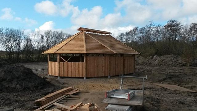 Et træhus med tag i konstruktion, omgivet af byggeplads og natur under en blå himmel.