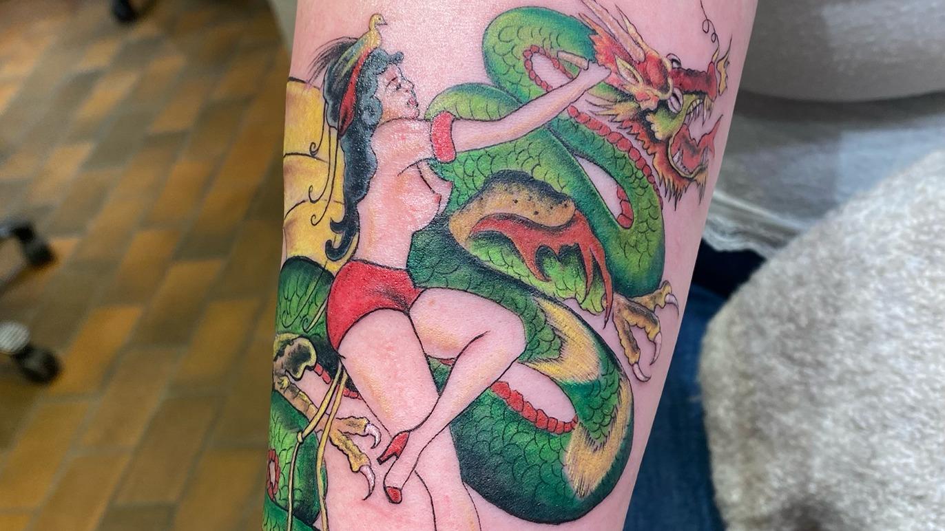 Farverig tatovering af en kvinde i rød badedragt, der kæmper mod en drage, med en grøn slange i baggrunden.