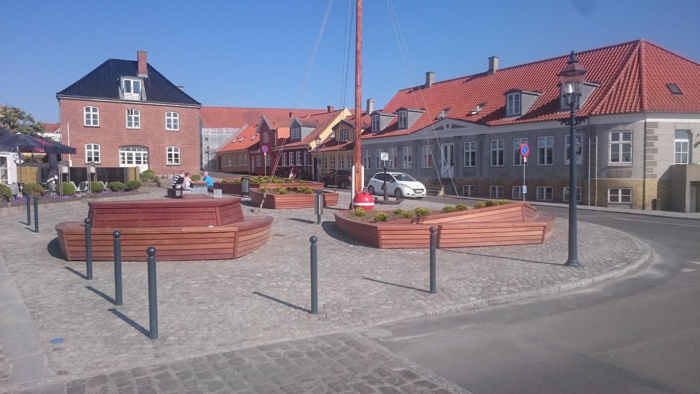 En solrig plads med træbede, brosten og bygninger i baggrunden, samt en bil parkeret ved siden af.