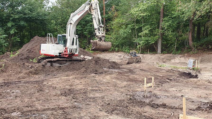 Gravemaskine arbejder på en byggeplads omgivet af træer og mudret jord.
