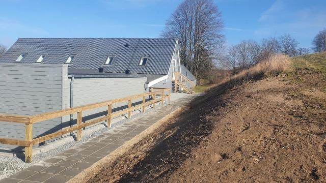 Nybygget hus med grå facade og skråning i forgrunden, omgivet af træer og blå himmel.
