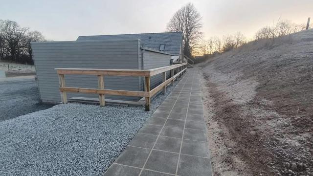 En grusbelagt sti ved en moderne bygning med trægelænder og en skråning i baggrunden ved solnedgang.