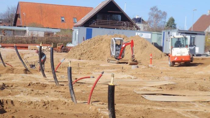 Byggeplads med gravemaskine, sandhøj og byggecontainere under konstruktion.