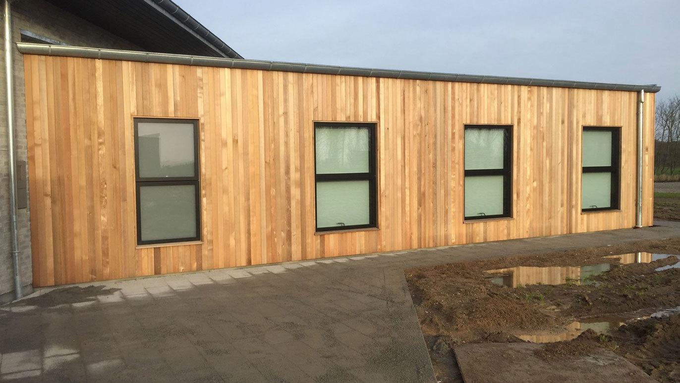 Moderne bygning med træfacade og store vinduer, beliggende på en grusvej.