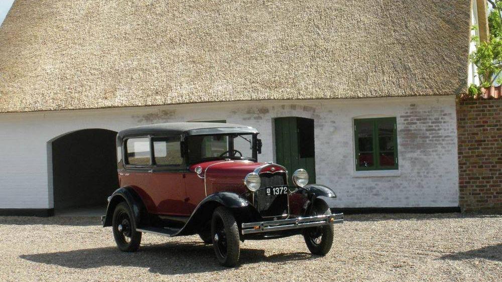 En gammel rød bil parkeret foran et stråtækt hus med hvide vægge.