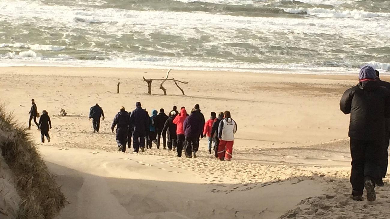 Gruppe mennesker går ned ad en sandstrand med bølger i baggrunden og en grå himmel.