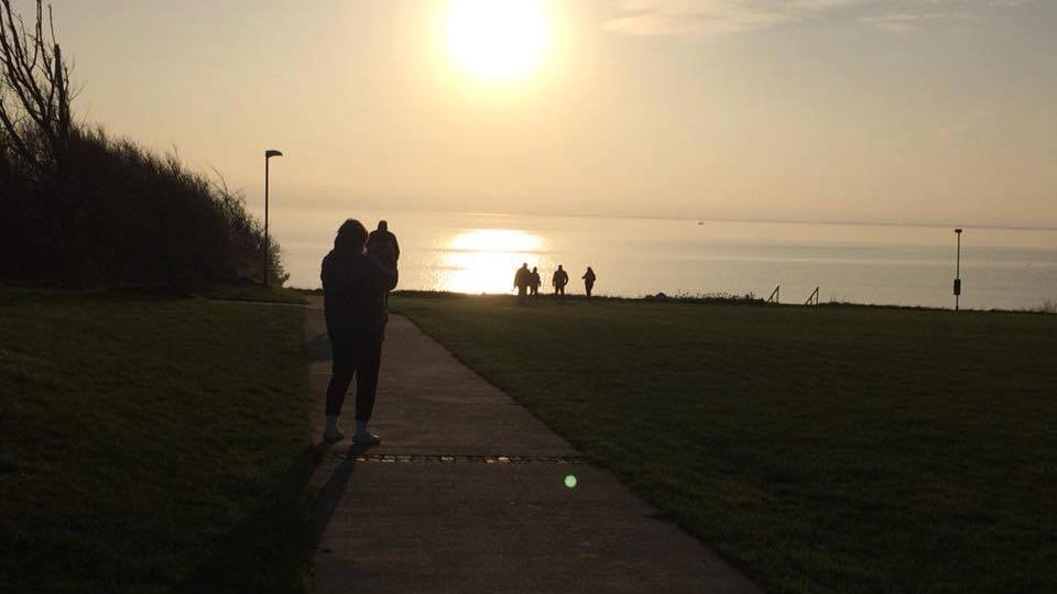 Solnedgang over vandet med mennesker, der går langs stien i en park.