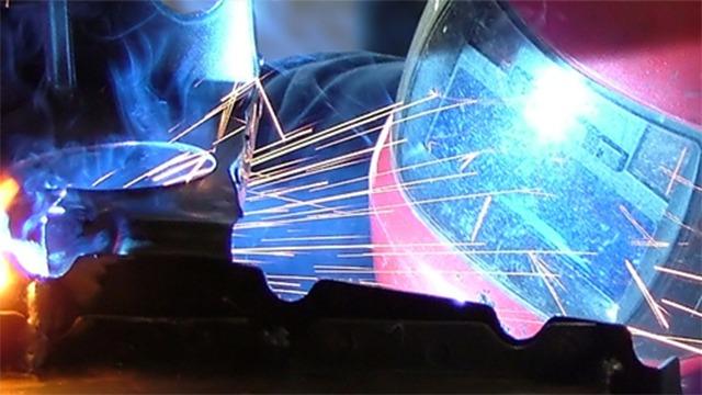 En svejser arbejder med en svejsemaskine, mens gnister flyver og lys reflekteres fra hjelmen.