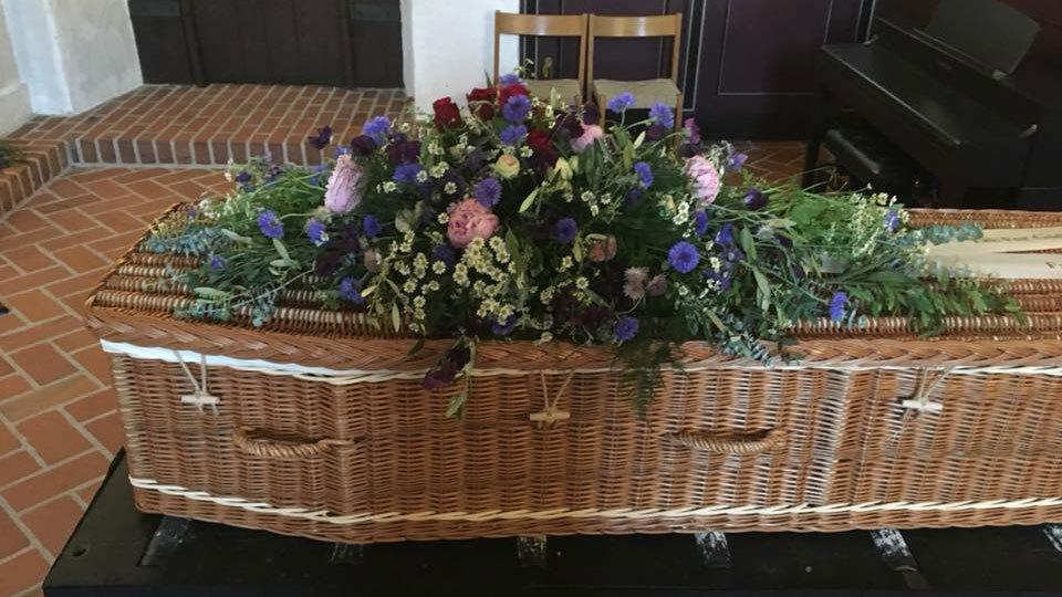 En flettet kiste dekoreret med farverige blomster og grønne planter i en kirke.