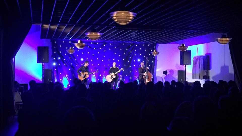 Tre musikere optræder på en scene med blå lys og stjerner, mens publikum ser på.