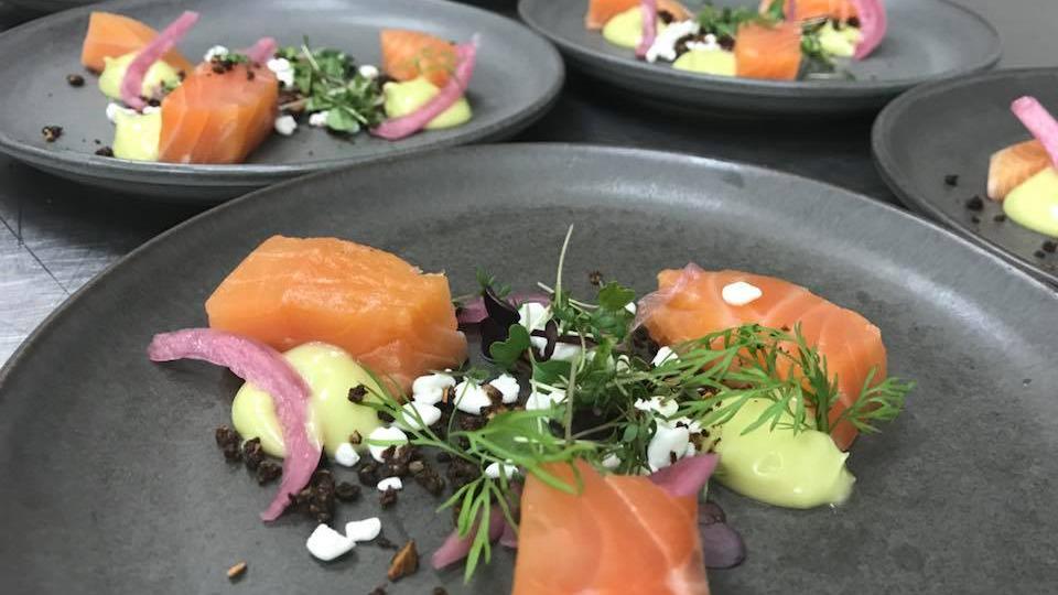 Lækker anretning med røget laks, cremet avocadomos, syltede rødløg og friske krydderurter på en mørk tallerken.