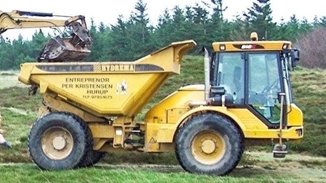 En gul dumper parkeret på en græsmark med skov i baggrunden. Maskinen er klar til at transportere jord.