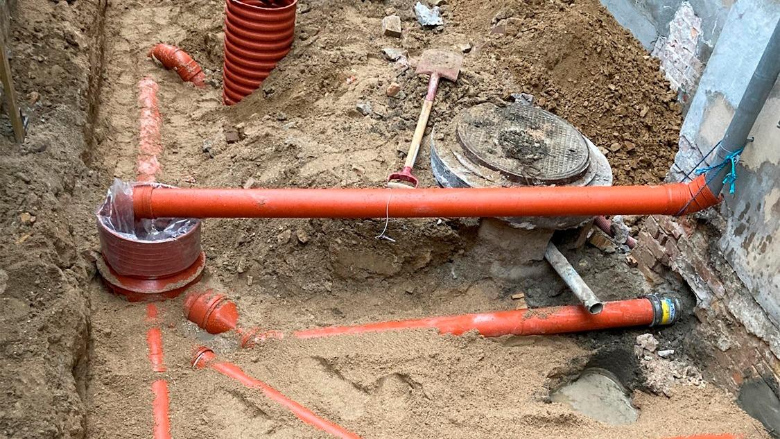 Rørinstallation i en grav med rød PVC-rør og værktøj til byggearbejde i sandet.