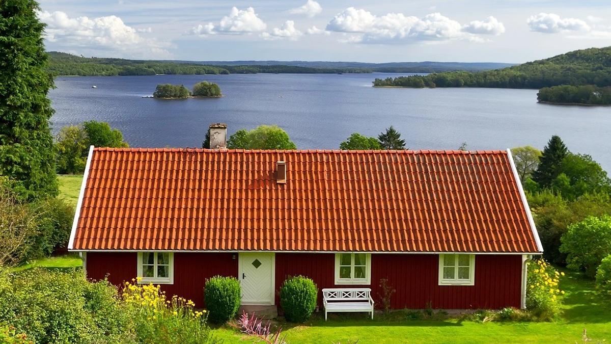 Et rødt hus med tag af teglsten ved en sø omgivet af grønne træer og bakker under en blå himmel.
