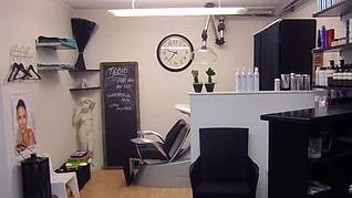 En frisørsalon med en sort stol, en væg med produkter, en klokke og en tavle med informationer.