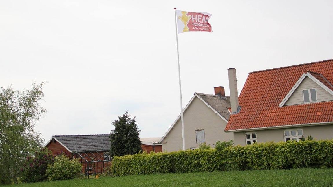 Hus med rød tag og flag, der blafrer i vinden, omgivet af grøn græsplæne og buske.