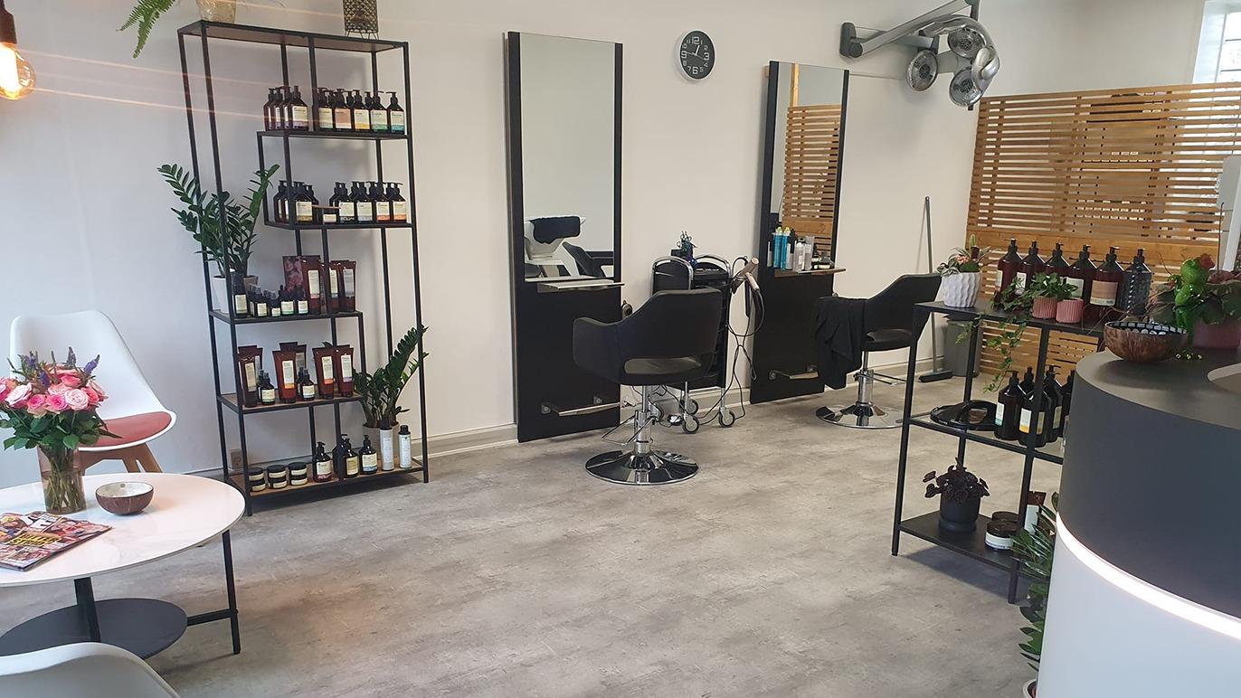 En moderne frisørsalon med spejle, stole, hylder med hårprodukter og en hyggelig siddeplads med blomster.