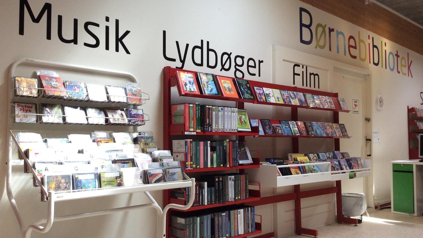 Børnebibliotek med hylder fyldt med musik, lydbøger og film i et lyst rum.