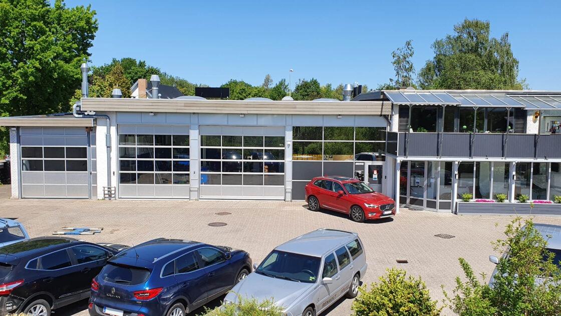 En moderne bygning med store vinduer, en rød bil parkeret foran og flere biler i parkeringspladsen.
