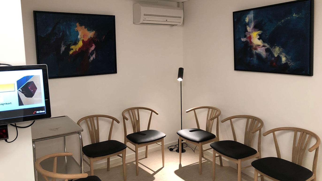 Et venteværelse med seks stole, en lampe og to farverige malerier på væggene. En skærm viser information.