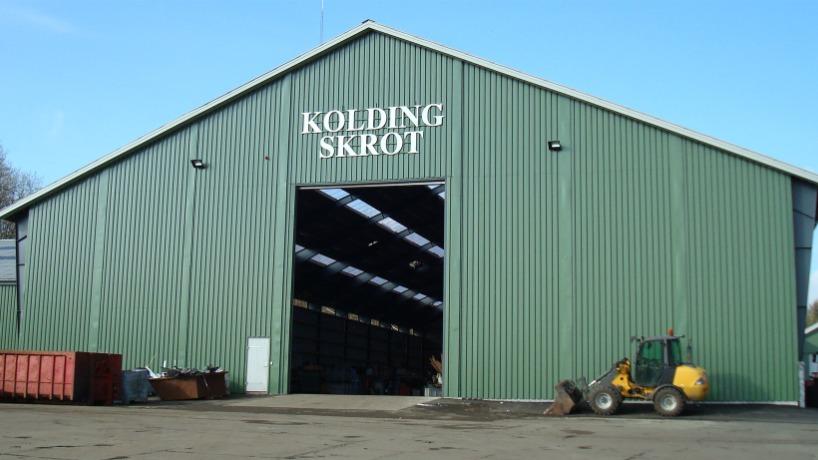 Grøn metalbygning med teksten "Kolding Skrot" over indgangen og en gule maskine foran.