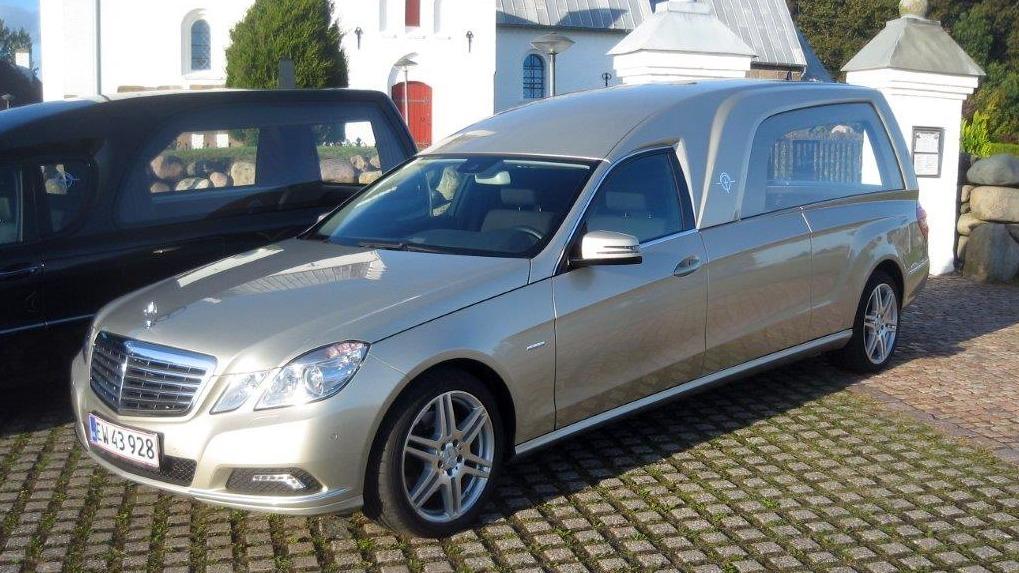En sølvfarvet ligvogn parkeret på en brostensbelagt plads ved en kirke.