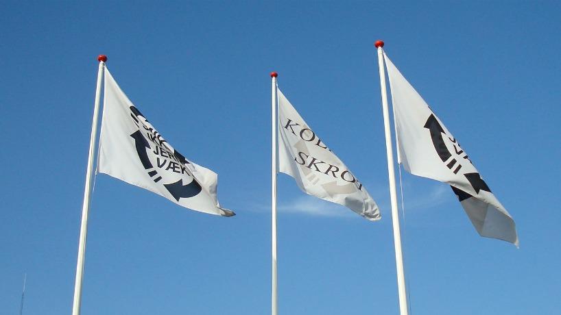 Tre hvide flag med sort tekst, der blafrer i vinden mod en klar blå himmel.