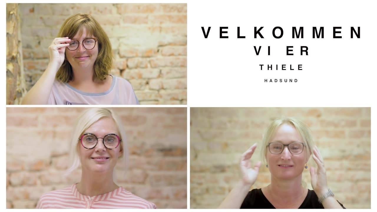 Tre kvinder med briller smiler og poserer foran en murstensvæg, med teksten "Velkommen vi er Thiele" i baggrunden.