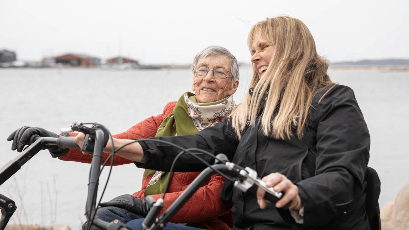 To kvinder på cykler ved vandet, smilende og nyder en hyggelig dag sammen.