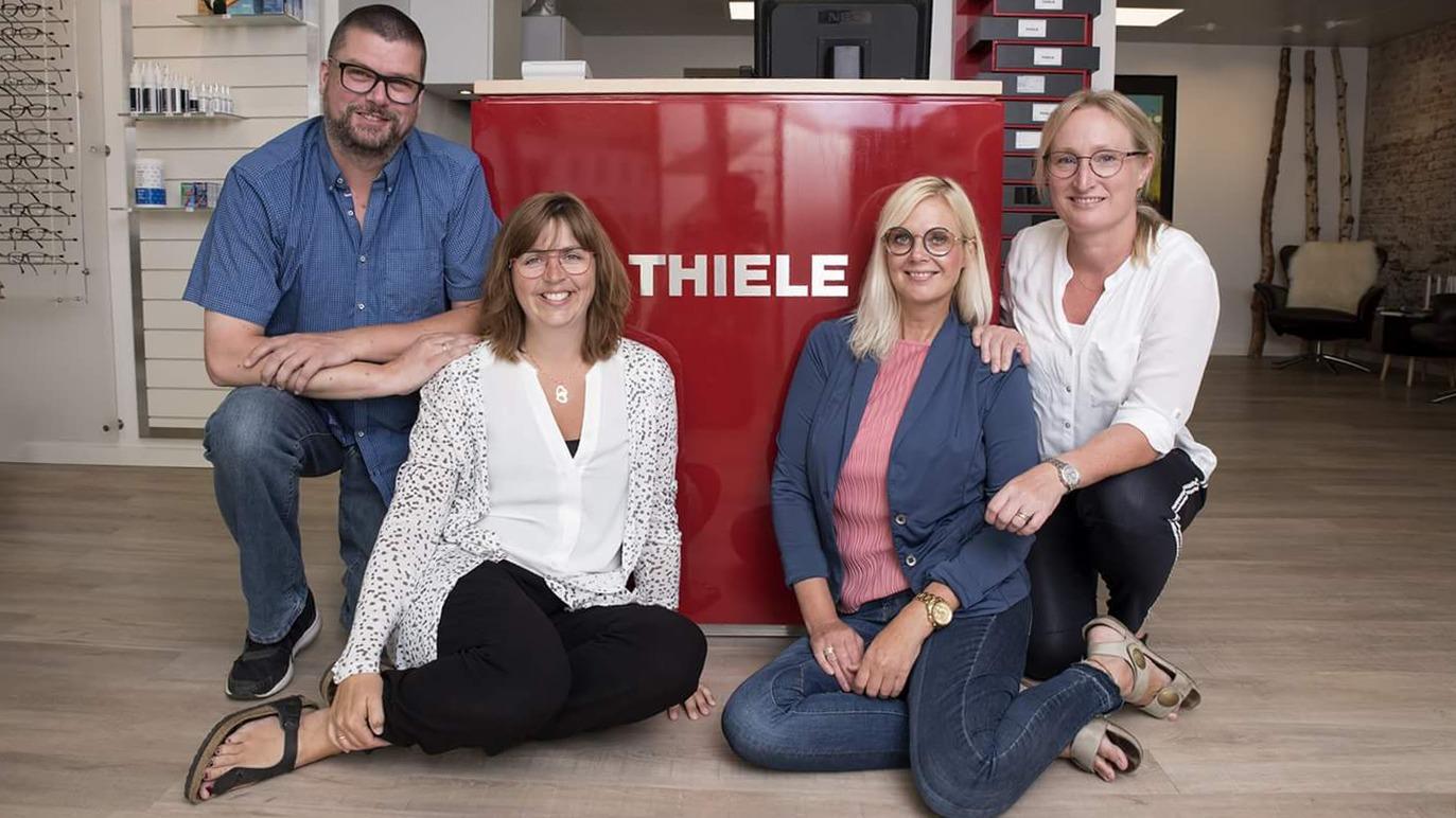 Fire personer poserer foran en rød disk med teksten "THIELE" i en moderne salon.