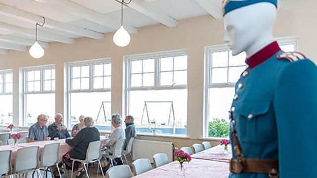 En mannequin i blå uniform står ved et bord, hvor ældre mennesker sidder og snakker i et lyst lokale.