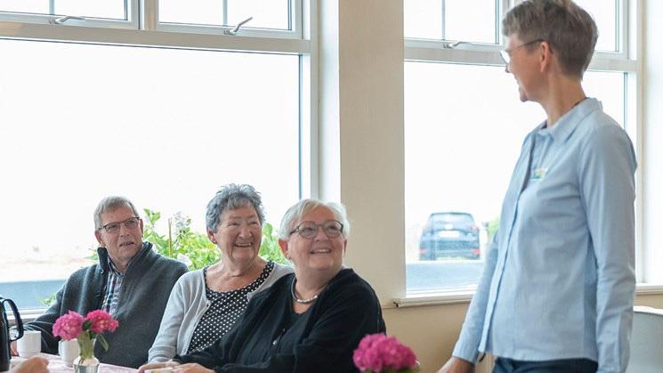 En kvinde står og taler til tre smilende personer ved et bord med blomster.