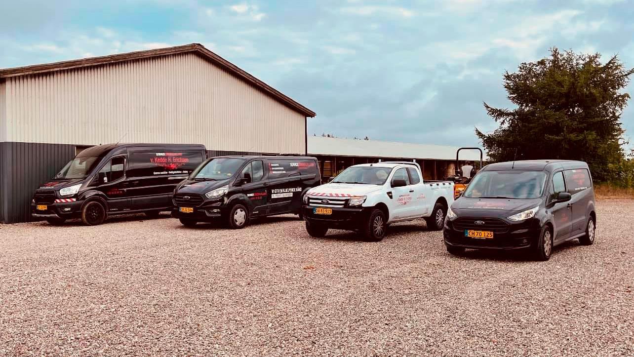 Flere varevogne parkeret foran en lagerbygning på en grusplads.