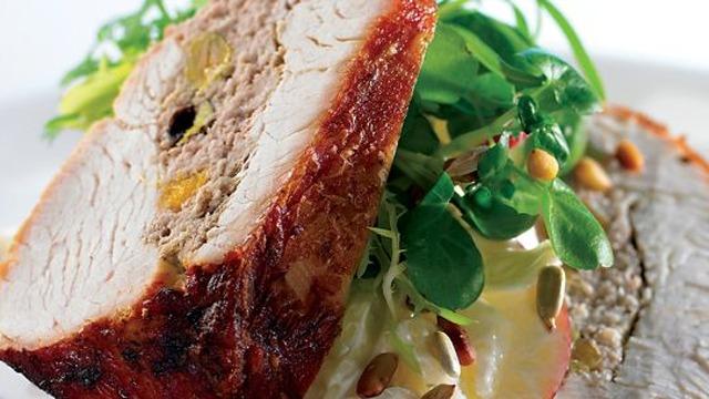 Lækkert stykke kød med fyld, serveret med friske salatblade og pinjekerner på en hvid tallerken.