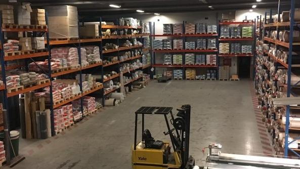 Lager med reoler fyldt med kasser og produkter, en gaffeltruck står klar til brug i midten af rummet.